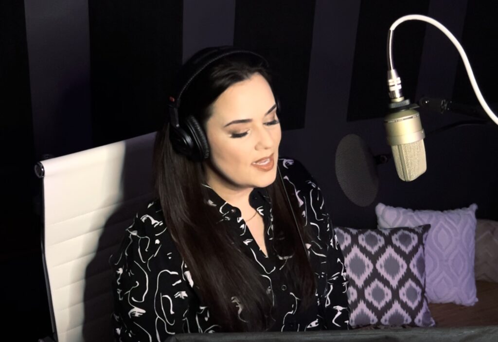 Voiceover artist Catherine Provenza wearing headphones sits in front of a microphone in a recording studio with pillows in the background.